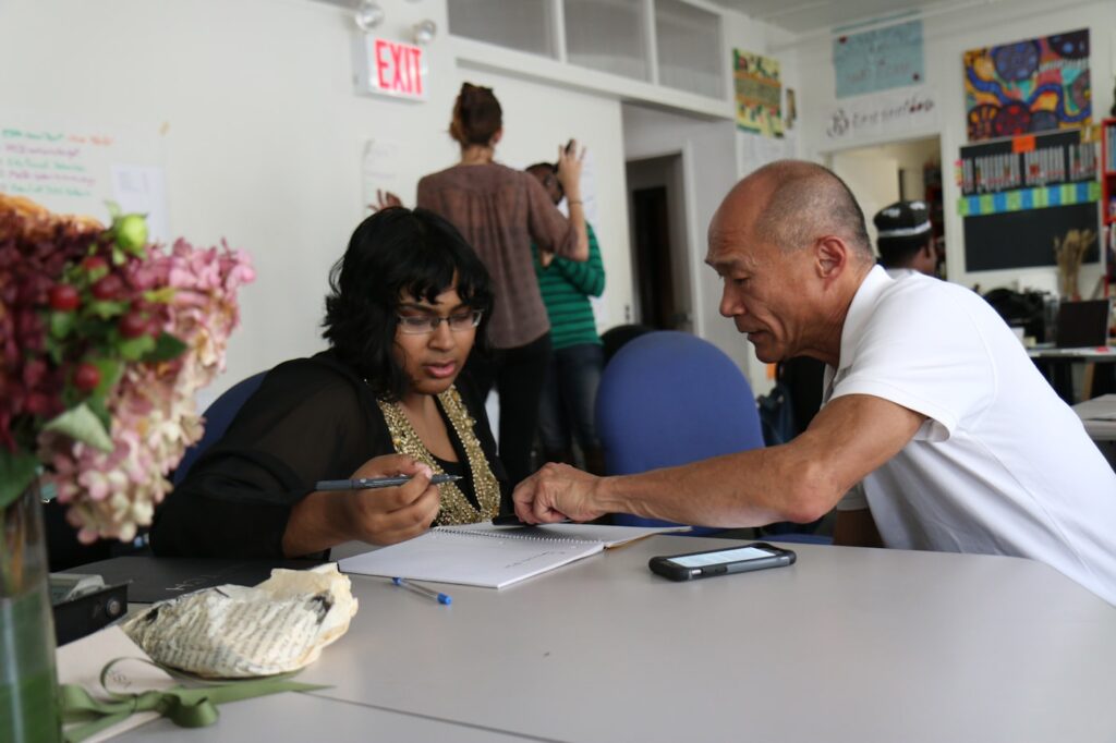 Older man mentoring a woman at a table, reviewing notes together, an example of contribution and mentoring after retirement